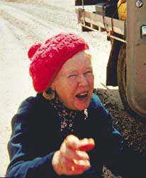 A woman wearing a red woollen hat with a pompom on top smiles, pointing her right hand at the camera. The tray of a motorised vehicle is partially visible in the background.