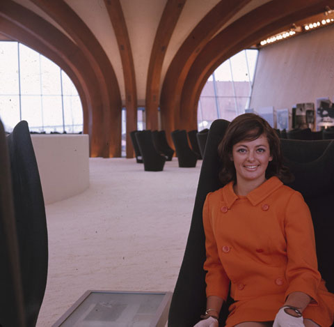 A woman dressed in an orange uniform and beige gloves sits on a dark green high-backed chair. Several similar chairs can be seen in the background.