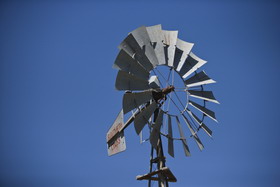 Top section of a windmill, set against a blue sky. Grey blades in a circular formation front a sail with 'SOUTHERN CROSS' painted in red.