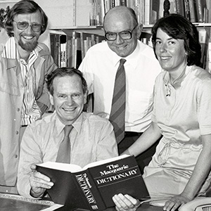 a black and white photo of four people inside an office. There is a bookcase behind them and the man seated is holding a Macquarie Dictionary