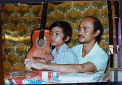 A man sits at a table with a young boy on his lap. Behind them a guitar is visible.