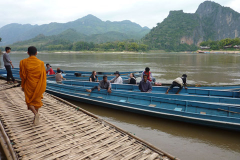 A man dressed in orange robes stands on platform of woven grass, beside a wide river. Various people sit in several blue wooden boats moored to the floating platform. Green mountains can been seen beyond the river.