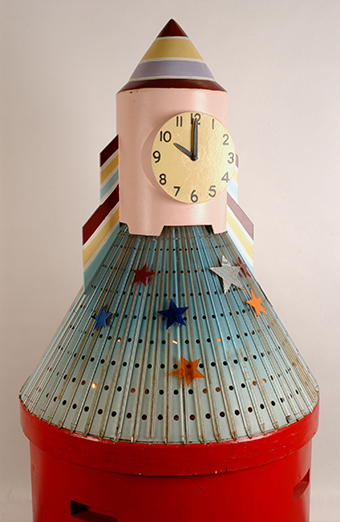 A large clock in the shape of a rocket with a red cylindrical wooden base with a light blue perforated metal skirt covered in vertical plastic rods, a light pink wooden trunk and painted striped cone top.