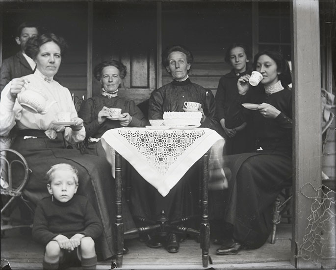 Group portrait of seven people posing on a veranda. Four women sit at a small wooden table. The women hold teacups and the woman at far left is also holding a teapot. At the foot of the table, sitting on the edge of the verandah is a young boy. Another young boy and a young girl stand behind the women at the table. On the table is a lace tablecloth, a teacup and a cake.