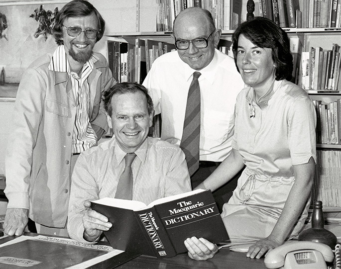 A black and white photo of four people inside an office. There is a bookcase behind them and the man seated is holding a Macquarie Dictionary