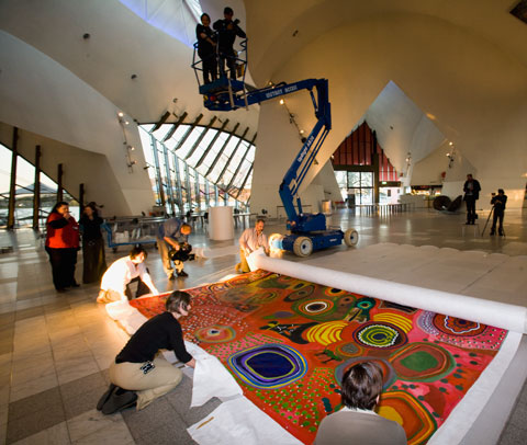 A colour photograph showing museum staff unrolling a large Indigenous painting on the floor of a museum hall. Three of the staff are closest to the camera, while a fourth is at one end of the rolled section of the painting. The visible section of the painting shows a brightly coloured background upon which have been painted numerous traditional patterns and forms. In the background of the photograph, two people watch the unrolling from the bucket of a high-lift crane. Another person is visible filming the unrolling and two others stand in the background with what appears to be camera equipment. One of them has a tripod with what appears to be a camera mounted on top.