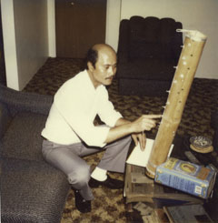 A man sits on a lounge. His right arm points to a bamboo musical instrument, which rests on a small table. The instrument has keys and strings down the side and an olive oil tin at the base.