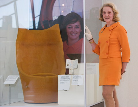 A woman wearing an orange top and skirt stands with her gloved right hand resting on a display case. An orange chair sits in the case, which includes a portrait of another woman at the rear.