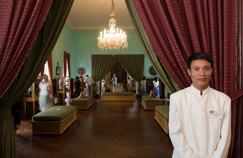 A man wearing a white coat stands to the right of a set of red and green curtains which frame a room with a lit chandeleir hanging from the ceiling and green upholstered seats along either side. Various people move about in the room.