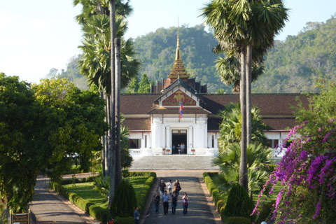 A regal building with a white facade, reddish brown roof and a gold spire rising above. A blue and red flag hangs over the entrance and an avenue of palm trees leads to the building. Manicured gardens are visible to either side and a treed hill appears behind the building.