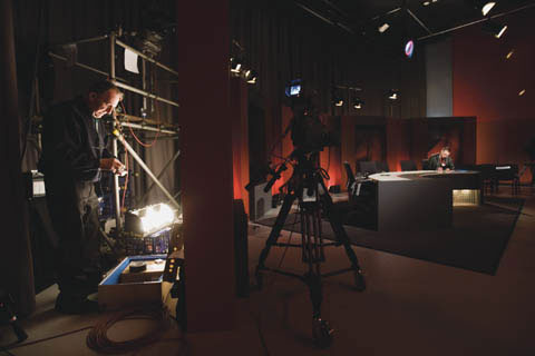 Behind-the-scenes image showing a television set in a studio, with a camera in the centre of the image and a man seated at a semi-circular shaped table at the right. A man stands on the left of the image beside scaffolding, cabling and technical gear.