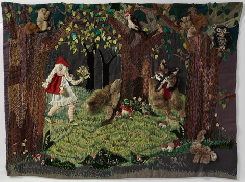 Wall hanging depicting Little Red Riding Hood walking through a forest. A wolf appears from behind a tree, with his red tongue hanging out the side of his mouth. A variety of fabrics including felt and fur have been sewn onto a grey blanket to form the scene, which also shows an owl, a bird, a squirrel and a rabbit. Some of the elements are padded using a technique called stumping, and they have a three-dimensional appearance.