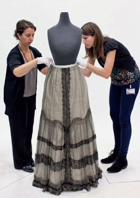 Two women stand either side and slightly to the rear of a mannequin. The mannequin's torso has neither arms nor a head and is finished in a dark grey material. The women, both wearing short white gloves, lean in to adjust the waistband of a skirt, which covers the lower half of the mannequin. The long skirt is mainly light grey in colour, with black lace down the centre, around the hem and in three vertical bands from about knee to hem.