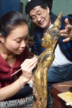 A woman leans in to apply a fine brush to a small gold sculpture. A man observes her work and supports the rear of the statue's head.