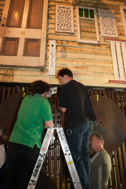 A colour photograph of two men standin on a ladder against a wall. There is another man to the lower right and a person on the lower left of the photograph.