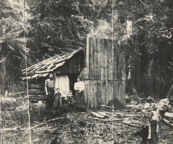 Family shot posing outside a timber hut with smoke rising from the chimney and logs lying on the ground.
