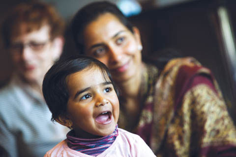 A young girl, singing, is in focus at the centre of the shot, with her mother, smiling, and slightly out of focus in the background.