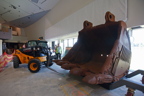 A loader with two forks extending from the front sits to the left of a large metal bucket which towers over two people and has five metal teeth protruding from the bottom.