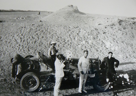 Black and white image showing four men standing beside an open-topped car in the desert. A sand dune rises above the men in the background.