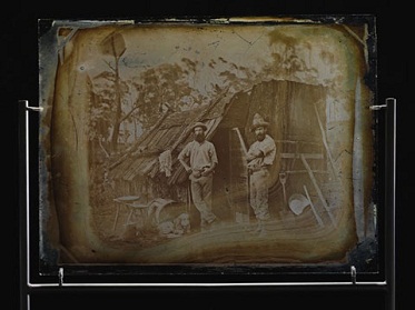 A piece of glass showing a photograph of two men standing outside a bark slab hut.