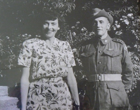 A black and white photograph of a man and a woman. The woman stands at the left of the photo, wearing a dress with a floral print. She has dark hair, and smiles. The man stands at the right of the photo, wearing a Second World War-era army uniform and slouch hat. He has his hands behind his back. Behind them both are large flowering shrubs and bushes.
