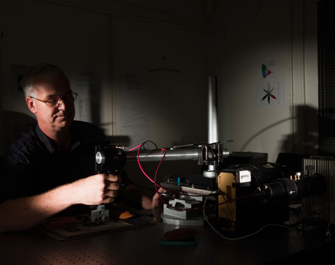 A colour photograph of a museum technician using specialist equipment to test the light sensitivity of an object. The technician sits at the left of the photograph, facing toward the right. On a table in front of him is the equipment, which appears to consist of a horizontal arm that is positioned over the object which is held in place on a special jig. Several wires are connected to the equipment. The photograph has dramatic lighting that has thrown some areas of it into deep shadow.