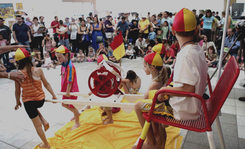 A crowd of spectators in the Museum's Hall watch a group of children pretending to be lifesavers. The children are wearing red and yellow swimcaps and carrying a surf-reel.