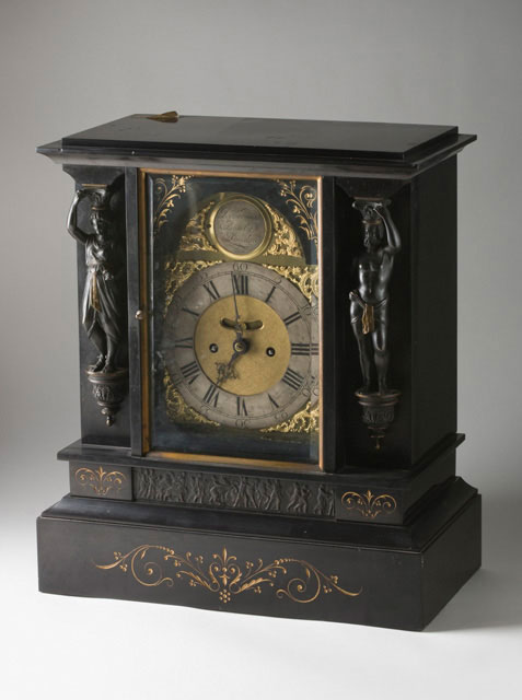 A mantelpiece clock, showing the face and one of the sides. The clock is made from dark material and has carved decorations.
