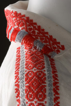 Side view showing a section of white linen shirt with the collar and sleeve embroidered in orange and grey thread. The collar is tasellated with the same thread.