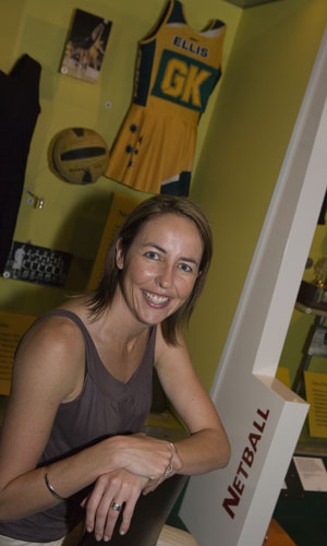 Liz Ellis sits in front of display case showing imagaes, netball and green and gold 'Ellis GK' uniform.