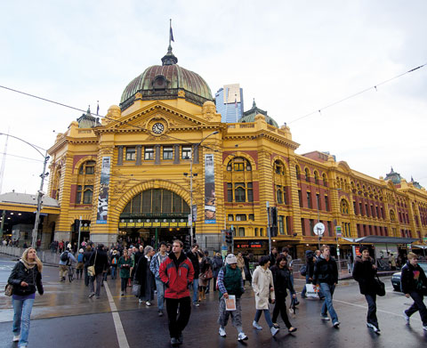 A crowd of people walks across a paved road outside Flinders Street Station, an Edwardian baroque building with a golden-coloured and red brick facade. Tram lines extend in various directions above the road.