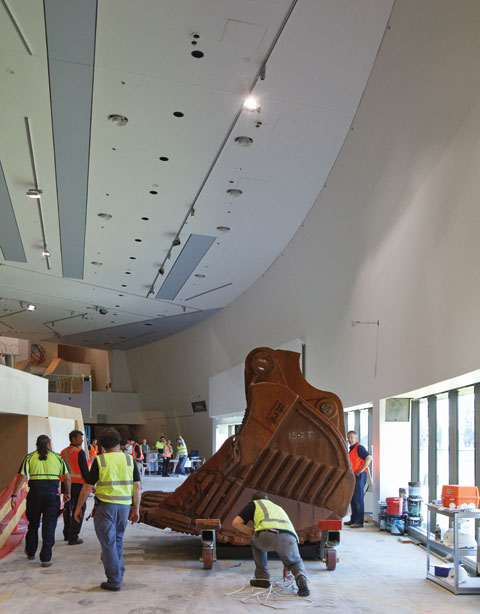 Side view of a rusty, concave-shaped iron bucket from a mining excavator. '15.2T' is spray-painted in white on the side. The bucket rests on a wheeled trolley and sits in a large, caverous gallery. Several workers in flourescent-coloured vests stand near the bucket and others gather in the distance.