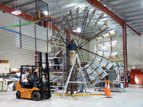 Rear view of a large windmill turntable and tail. A forklift holds the corrugated iron tail in place while two men work at height to attach it to the circular turntable.