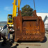 A view of a large mining excavator bucket being lifted off a semi-trailer. The bucket hangs a few centimetres off the ground, with the back of it facing the camera. Beyond the bucket can be seen the large yellow crane that has lifted the bucket off the semi-trailer. In the left foreground of the image stand two men and a woman, with their backs to the camera. In the middle of the image two men can be seen preparing to place large pieces of timber under the bucket. In the right background can be seen part of what appears to be a large white industrial shed. At the far left of the image can be seen part of some other mining equipment which is on the semi-trailer. Part of the sky is visible beyond the activity in the foreground. A few clouds are visible in the far left background.