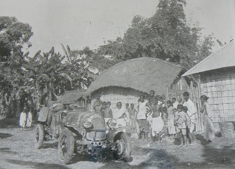Black and white photo showing an open-topped car in a village. A group of about 20 people stands to the right and several grass walled buildings and trees form a backdrop.