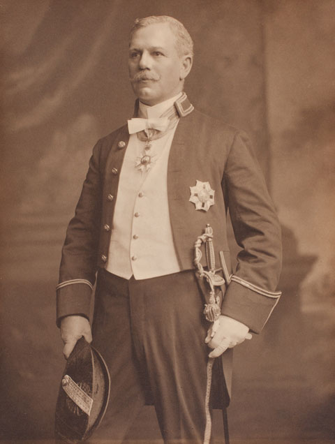 Black and white photograph of a man wearing a ceremonial dress uniform. The man wears ceremonial regalia and holds a bicorn hat in his left hand and a sword in his right.