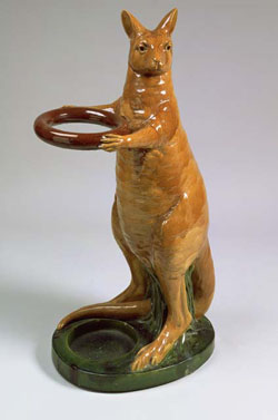 Ochre-glazed stoneware kangaroo, standing on a base. The kangaroo's paws extend around a ring and its feet and tail straddle a circular recess in the base.