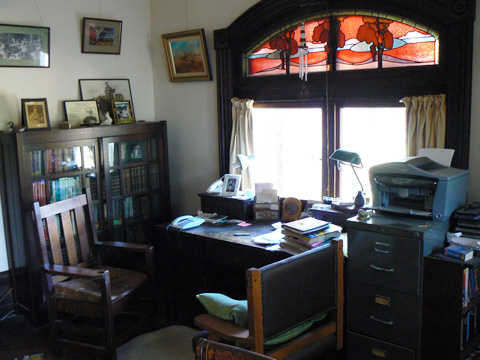 A room with a table, bookcase, filing cabinet and two chairs. The table faces a window that has a red, blue and purple stained glass feature above it. On the table are books, boxes, a tall lamp, a modern keypad telephone and loose papers. There are photographs on the bookcase and paintings hung on one wall.