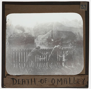 Black and white image of three men crouching behind a fence. The man at centre is reeling backwards. A building burns in the distance. 'DEATH OF O'MALLEY' is handwritten at the bottom of the slide.