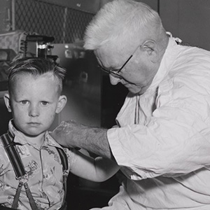 Interior black and white photo of a late middle-aged man in white coat administering injection to the upper arm of a boy, about five