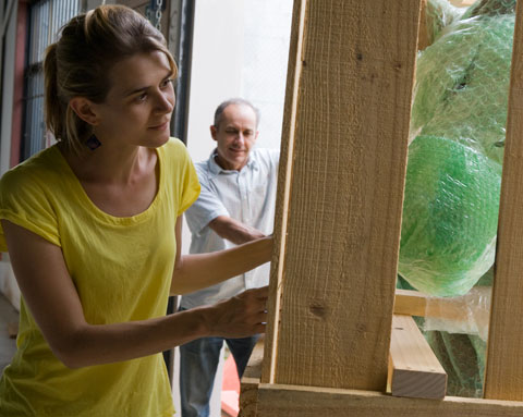 A colour photograph of two museum staff members preparing to unpack an exhibit. One, closest to the camera is closely inspecting the packing crate that contains the exhibit. The large wooden frame of the packing crate is visible at the right side of the photograph.