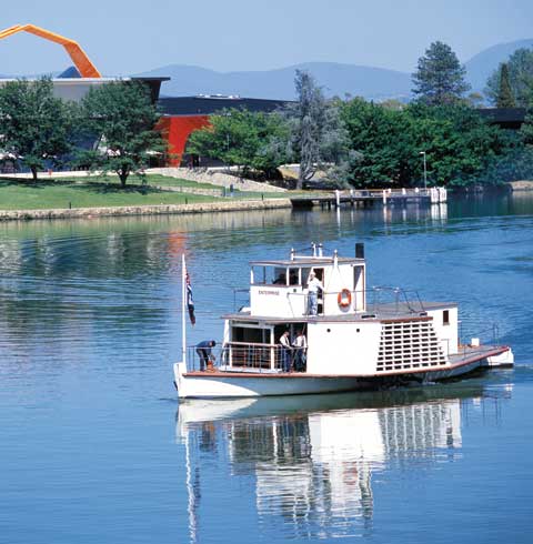 The wooden paddle steamer 'Enterprise' on Lake Burley Griffin, with the National Museum in the background. Four crew members stand on the steamer's deck.
