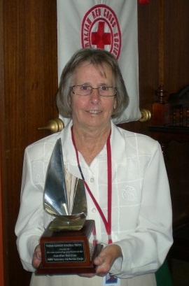 A woman stands at the centre of the image holding a trophy at her waist. The trophy has a curved metal feature on top of a wooden base, with a plaque reading 'YVONNE KENNEDY MEMORIAL TROPHY / Awarded for / the most outstanding contribution to the / Australian Red Cross / NSW Voluntary Aid Service Corps'. A Red Cross banner is partially visible in the background.
