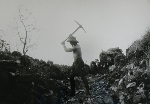 Black and white photo showing a the side view of a topless man wearing a pith helmet, with an axe raised above his right shoulder. He stands down a rocky gorge. An open-topped car sits at the top of the gorge.