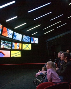 Colour photograph showing five people sitting at the right of a darkened room, looking at three rows of screens on the left. The nine screens show images of various colourful architectural details. Narrow slits of light are visible on the roof of the theatre.