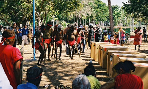 Community members and Aboriginal men dressed in red loin cloths gather around boxes containing remains
