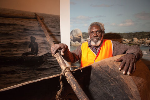 Colour photograph of a man standing behind a partially visible dugout canoe. Images of a canoe and a landscape are partially visible in the background.