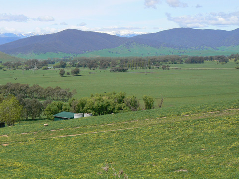 A colour photograph of a broad valley. In the foreground is a slope, and a building partially visible at the bottom of the slope. Beyond the building is an open plain and some trees at the far edge of the plain. In the background a mountain range is visible. In the far distance are some snow-covered mountain peaks. Some scattered clouds are visible in the sky.