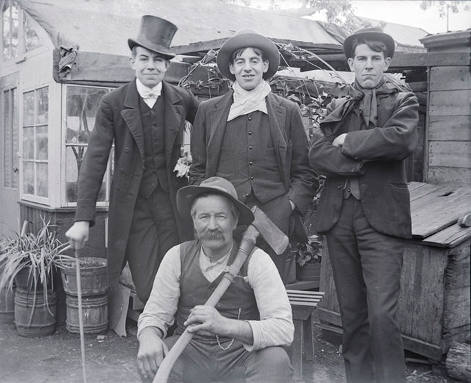 Group portrait of four men posing in front of a dwelling. Three young men stand at rear, wearing dark suits and hats. The man far left leans on a cane. The other two wear scarves. The fourth man, who is older, squats at the front, wearing a vest without a jacket and holding an axe, which rests on his shoulder.