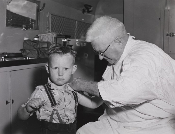 Interior black and white photo of a late middle-aged man in white coat administering injection to the upper arm of a boy, about five